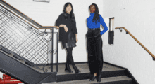 A woman wearing all black, including a leather skirt, combat boots and a cardigan, stands next to another woman wearing black dress pants, a blue turtleneck and black patent mules.