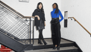 A woman wearing all black, including a leather skirt, combat boots and a cardigan, stands next to another woman wearing black dress pants, a blue turtleneck and black patent mules.