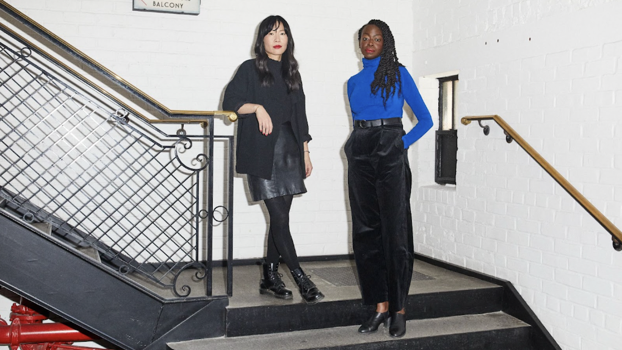 A woman wearing all black, including a leather skirt, combat boots and a cardigan, stands next to another woman wearing black dress pants, a blue turtleneck and black patent mules.