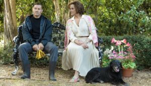 A man wearing all black and a woman wearing all white sit on an iron bench in a garden. A black dog sits at the woman's feet.