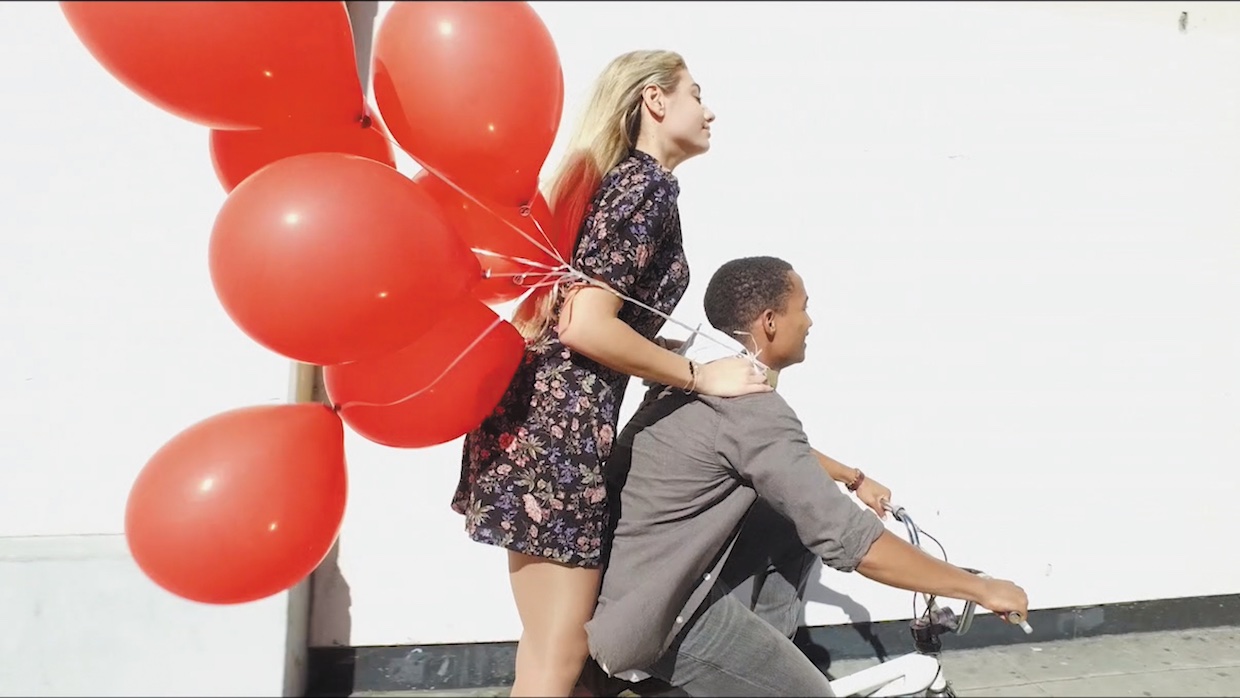 A woman with blond hair and a floral sundress rides standing on the back of a young Black boy's bike, a flurry of balloons flying behind them.