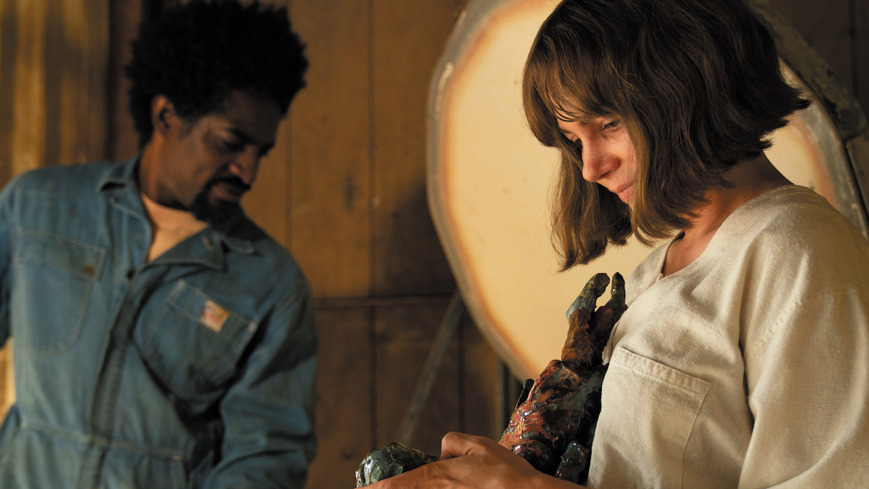 A woman with a dirty blond bob haircut holds two ceramic figures that have recently been fired in the kiln; a man wearing a utility jumpsuit unloads other kiln creations in the background.