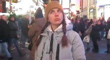 A young woman wears a gray parka and tan beanie and stands in the hustle and bustle of Times Square.