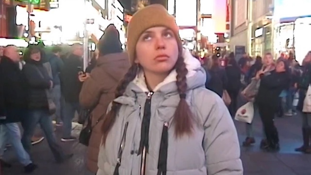 A young woman wears a gray parka and tan beanie and stands in the hustle and bustle of Times Square.