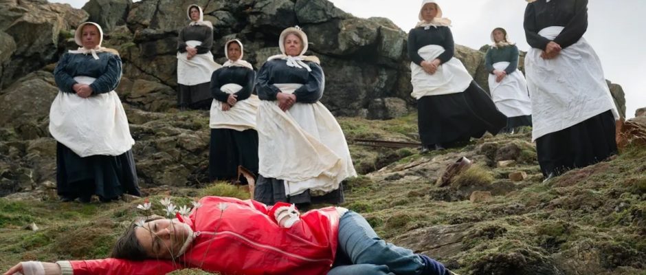 A woman in a red raincoat and blue jeans lies on her side on a rocky moor; she is surrounded by foreboding Puritan women.