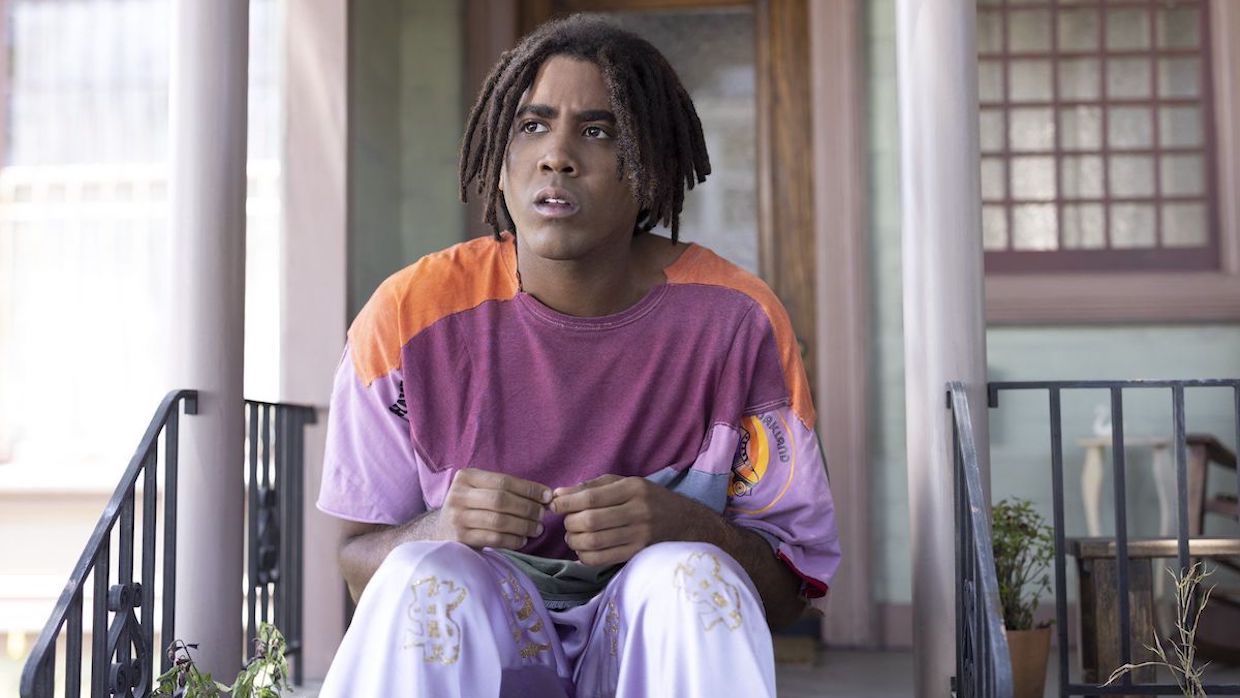 A tall Black young man sits on a stoop, wearing a purple and orange colorblocked shirt and sateen lavender pants.