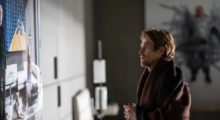 Willem Dafoe appears disheveled as he stares at art hanging on a wall, a blanket is draped over his shoulders and he holds a mug in his hand.