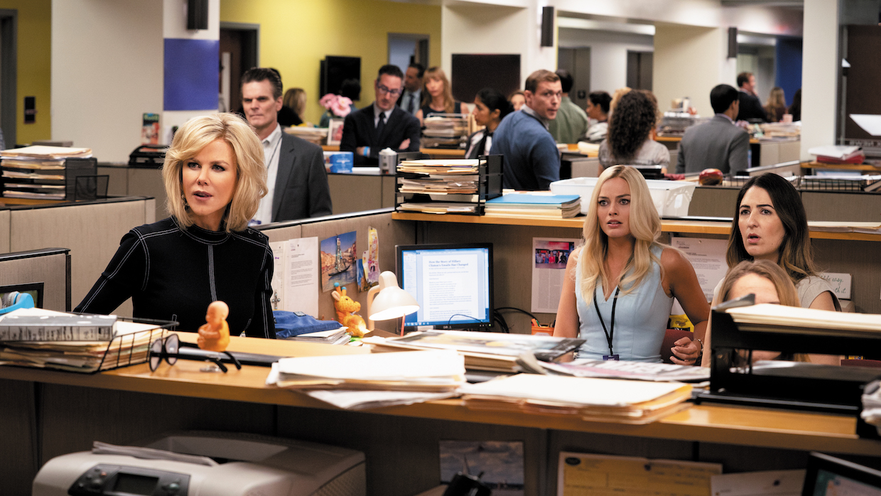 Three women, two blonds and a brunette, sit at their desks in a hectic newsroom.