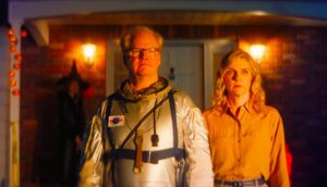 A middle-aged man wears an astronaut suit and stands next to a younger woman wearing a salmon colored button-up shirt.
