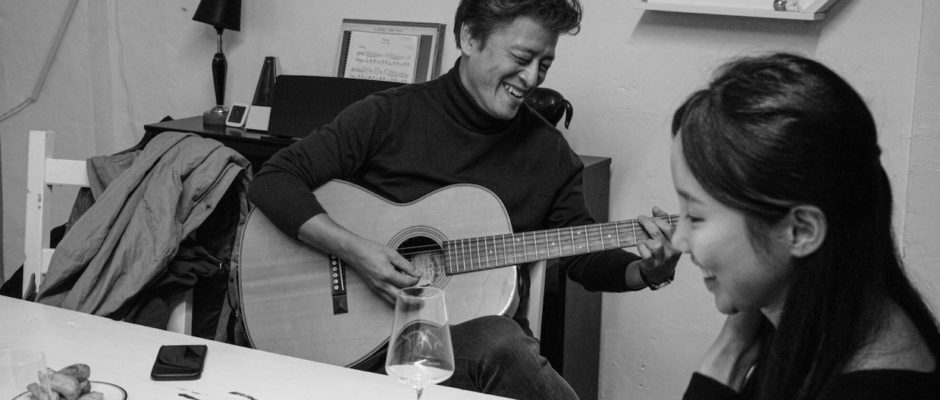A man wearing a black turtleneck plays guitar for a young woman who looks off into the distance.