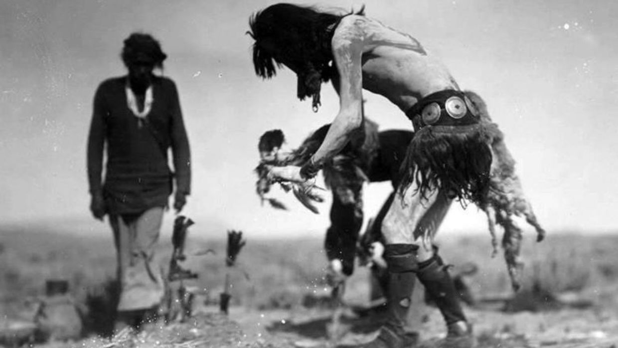 A black and white image of one shirtless man crouching down as another, out of focus man in more polished attire lingers in the background.