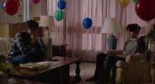Two brown-haired teenage boys sit in a living room. Despite the space being decorated with balloons and bright pink curtains, they both look morose.