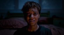 A young Black woman with cropped short hair sits up in a dark room, her eyes widen as she gazes at something that clearly terrifies her.