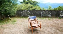 An empty wooden chair sits in a yard, the grass barely grows and is surrounded by a stone wall.
