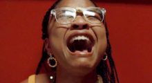 A Black teenage girl with braided hair, delicate hoop earrings and cracked clear glasses closes her eyes and screams.