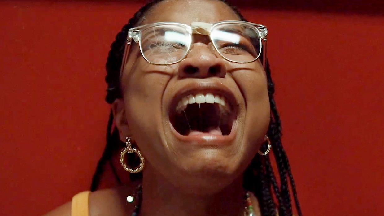 A Black teenage girl with braided hair, delicate hoop earrings and cracked clear glasses closes her eyes and screams.
