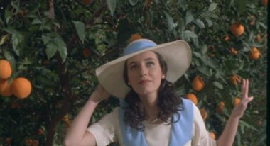 A woman with shoulder-length, wavy brown hair stands amid an orange grove. She wears a wide-brimmed hat with a blue ribbon that matches her dress.