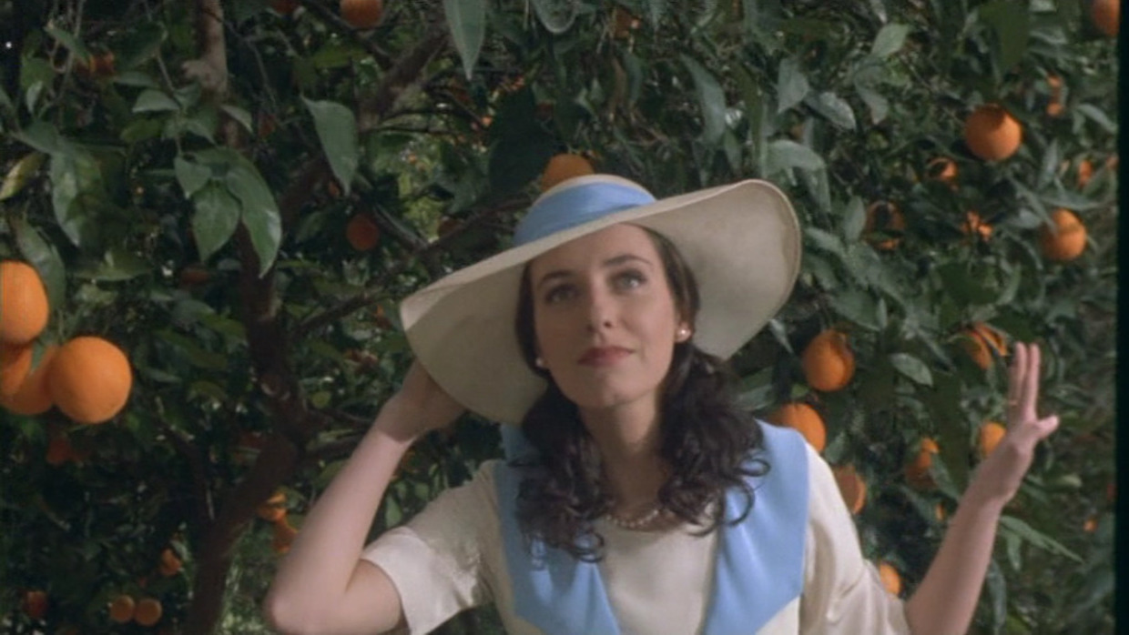 A woman with shoulder-length, wavy brown hair stands amid an orange grove. She wears a wide-brimmed hat with a blue ribbon that matches her dress.
