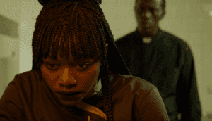 A close-up shot of a Black woman with braided hair. She leans over a counter while a priest looms over her from behind.