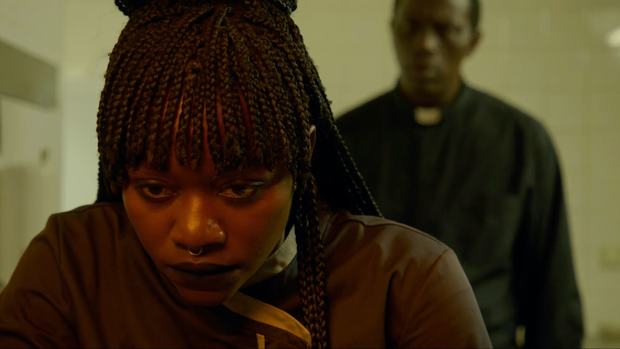 A close-up shot of a Black woman with braided hair. She leans over a counter while a priest looms over her from behind.