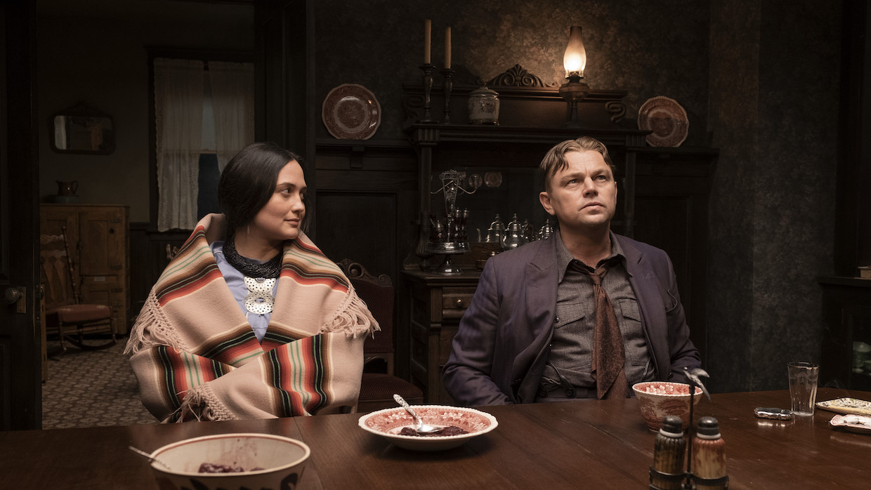 A woman wearing an orange and tan hand-woven shawl sits at a dining table next to a man in a dark-toned suit. She looks at him and he looks away into the distance. The room is like by a few stray gas lamps and the plates they have eaten from are empty.