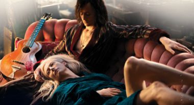 Two lovers sit on a burnt orange, antique velvet couch; the man has dark long hair and wears a dark robe with his chest exposed, the woman, who has platinum blond hair, lies across his lap, donning a teal robe and drapes her pale leg over the back of the tufted couch. An acoustic guitar sits by the man's side.