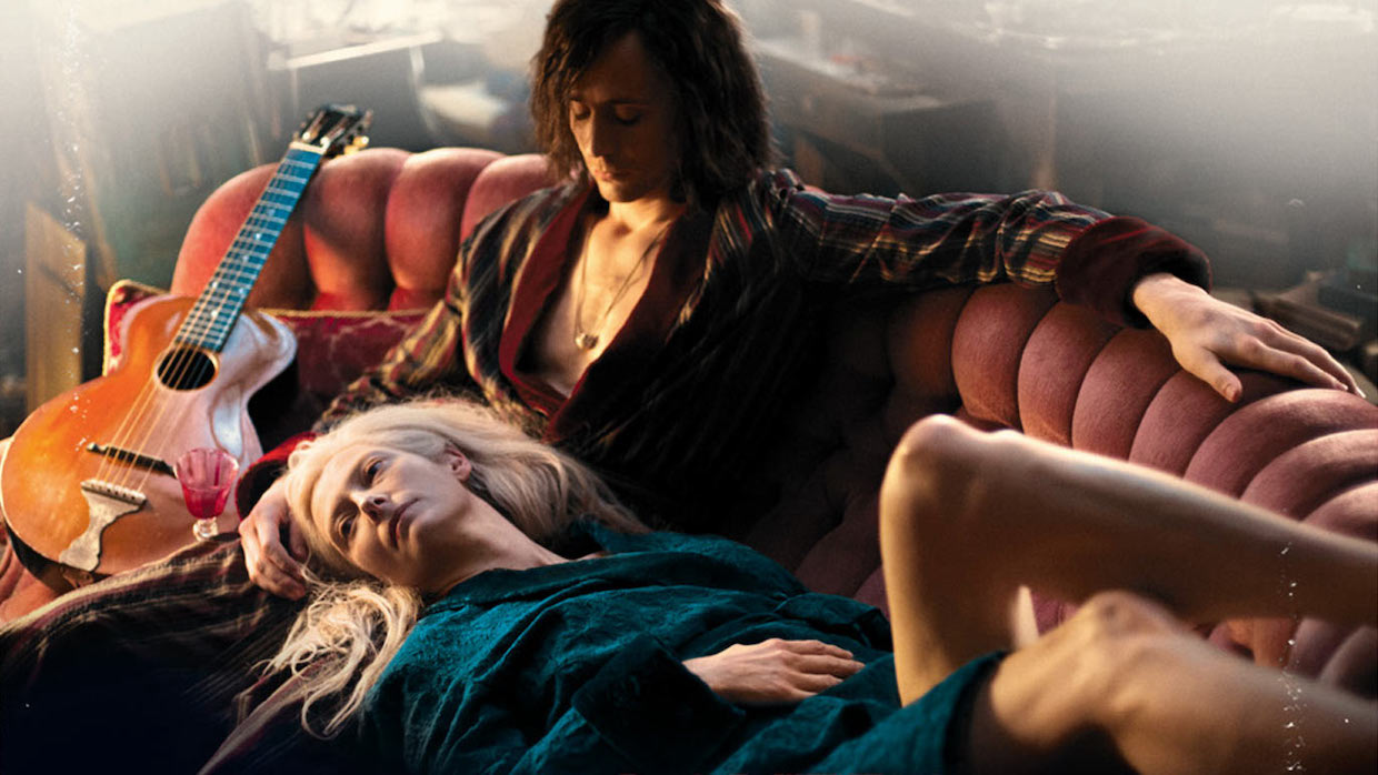 Two lovers sit on a burnt orange, antique velvet couch; the man has dark long hair and wears a dark robe with his chest exposed, the woman, who has platinum blond hair, lies across his lap, donning a teal robe and drapes her pale leg over the back of the tufted couch. An acoustic guitar sits by the man's side.