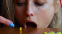 A blond woman with neon blue painted nails opens her mount near the edge of a table and rolls a bright green pill into her mouth.