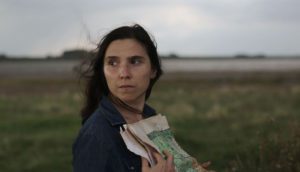 A woman stands in an open field, gray skies behind her, as she clutches a newspaper to her chest. The wind lightly blows her brown hair behind her.