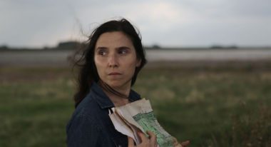 A woman stands in an open field, gray skies behind her, as she clutches a newspaper to her chest. The wind lightly blows her brown hair behind her.