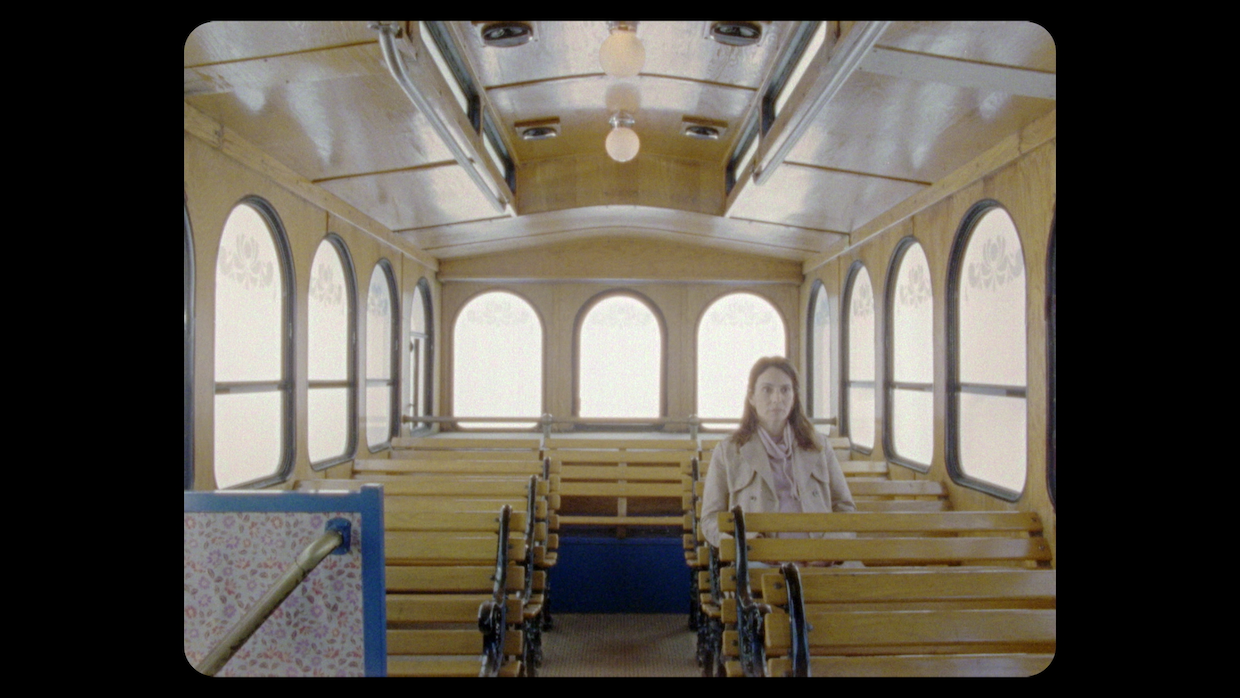 A woman sits on an empty old-fashioned trolley, she sits and stares straight ahead and the windows let in flat, white light.