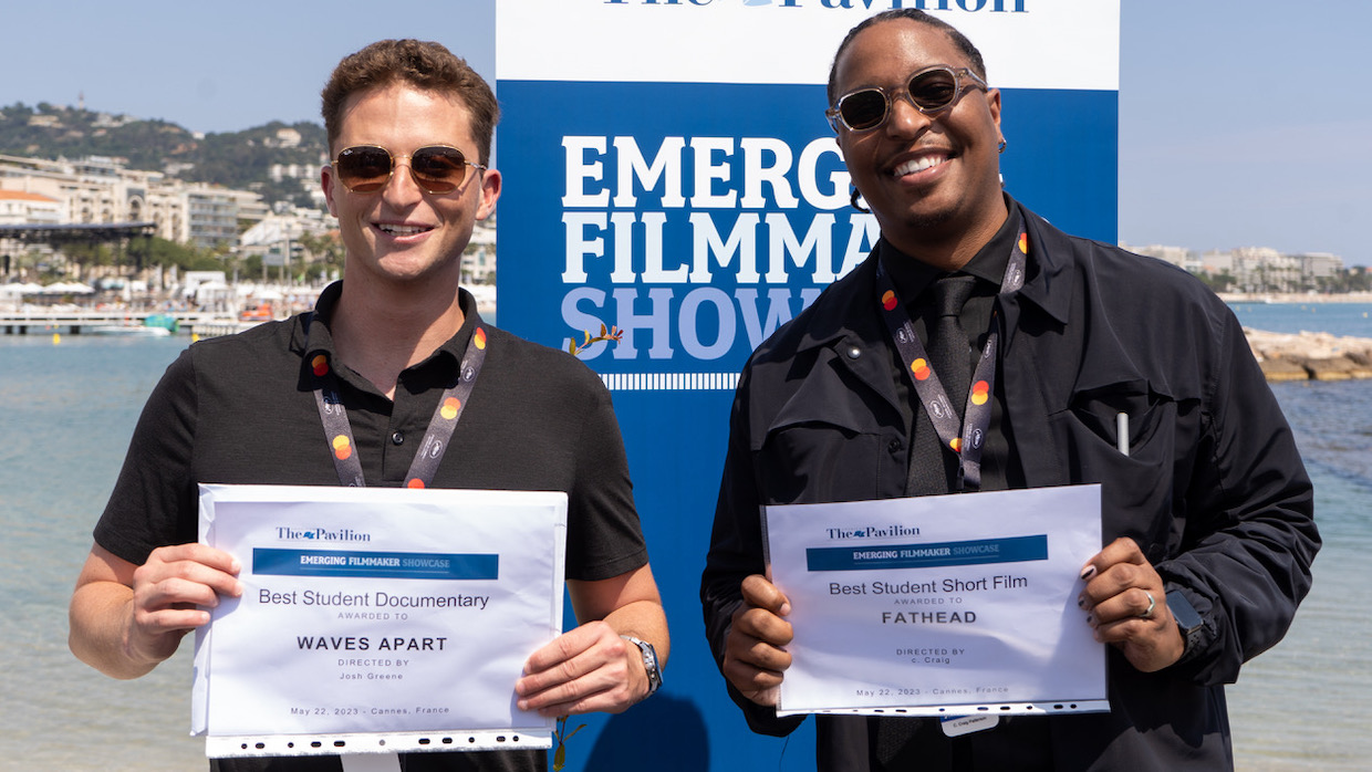 Two young men stand in front of the Emerging Filmmaker Showcase at Cannes and hold up paper certificates that show they've won.