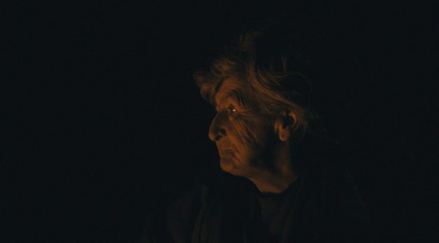 An elderly woman with white hair tied back in a bun stares to her right. She is illuminated only by the light of a fire that exists out of frame; her surroundings are pitch-black.