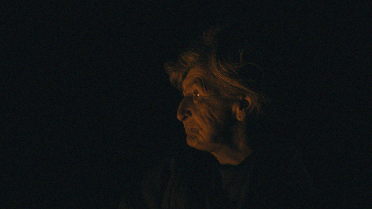 An elderly woman with white hair tied back in a bun stares to her right. She is illuminated only by the light of a fire that exists out of frame; her surroundings are pitch-black.