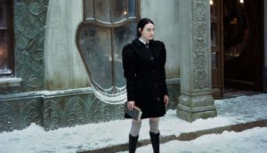 A woman with long black hair stands on a snowy sidewalk in front of a gray stone building with eccentric oblong windows. She wears a black peacoat with exaggerated shoulder pads and gray thigh-high socks with black knee-high boots.