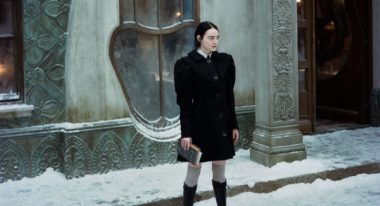 A woman with long black hair stands on a snowy sidewalk in front of a gray stone building with eccentric oblong windows. She wears a black peacoat with exaggerated shoulder pads and gray thigh-high socks with black knee-high boots.