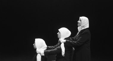 Three women wearing hijabs stand behind one another gripping the shoulders of the woman in front of them. They are organized in order of height, with the shortest woman at the front and tallest woman at the back in this stark black and white photo.