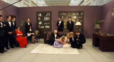 A cast dressed in sophisticated attire, including tuxedos and cocktail dresses, act out a dramatic scene where several people sit on the floor crying dramatically. Cameras, microphones and crew members are clearly visible.