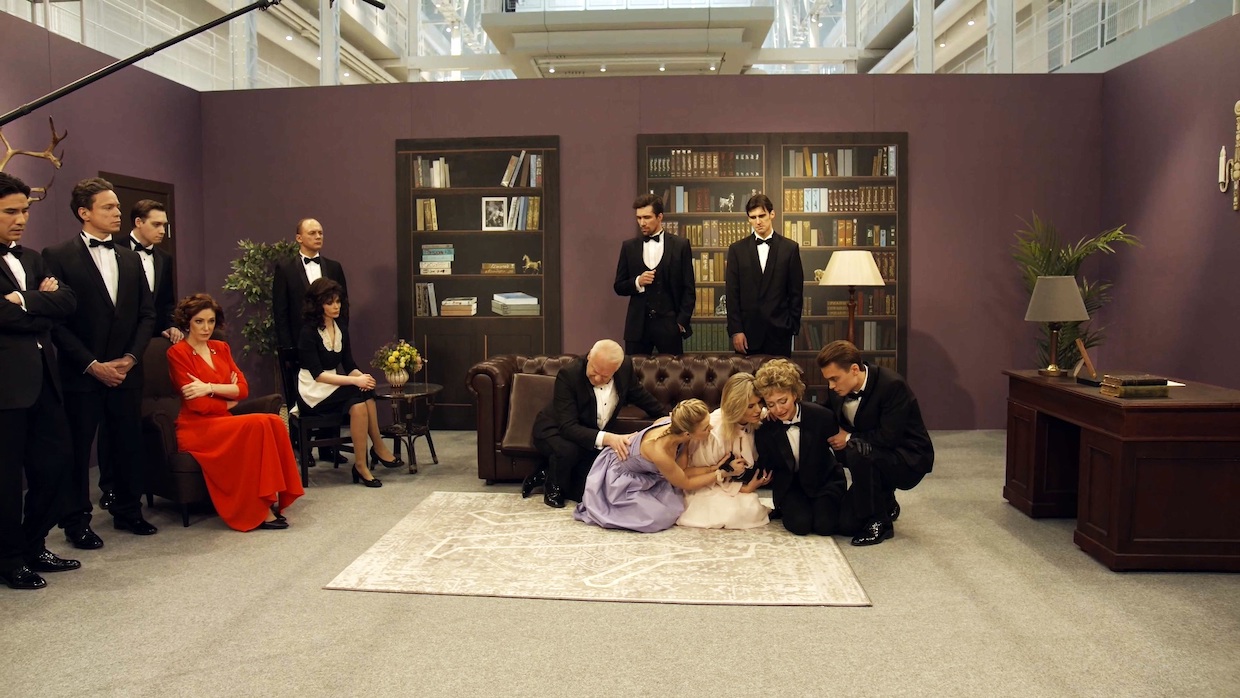 A cast dressed in sophisticated attire, including tuxedos and cocktail dresses, act out a dramatic scene where several people sit on the floor crying dramatically. Cameras, microphones and crew members are clearly visible.