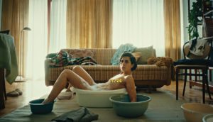 A woman sits nude in a plastic washtub in the middle of her living room. Her extremities also soak in similar plastic buckets.