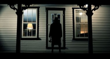 A shadowy figure holds and axe and stands in front of a house's door.