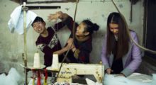 Three young Chinese people stand in front of a industrial sewing machine, they goof around and smile.