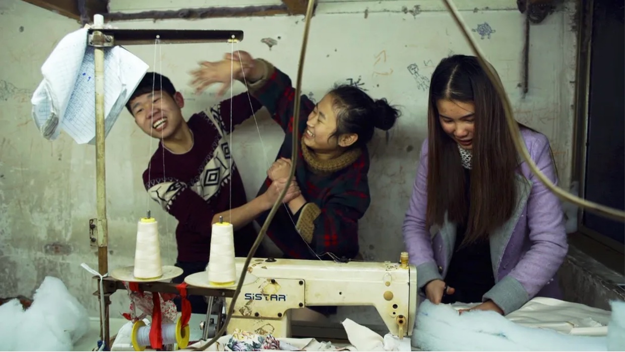 Three young Chinese people stand in front of a industrial sewing machine, they goof around and smile.