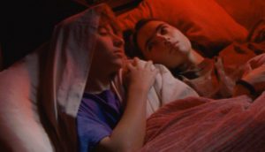 A teenage boy lays in bed with a girl. A red-tinted light fills the space.