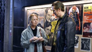 A woman with blond hair in a light blue coat holds a piece of paper and a pen in her hand as she looks as a tall man in a black leather jacket. They stand in front of a movie theater poster board.