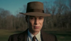 A '40s-era man with a brown fedora and matching suit stands and looks contemplatively.