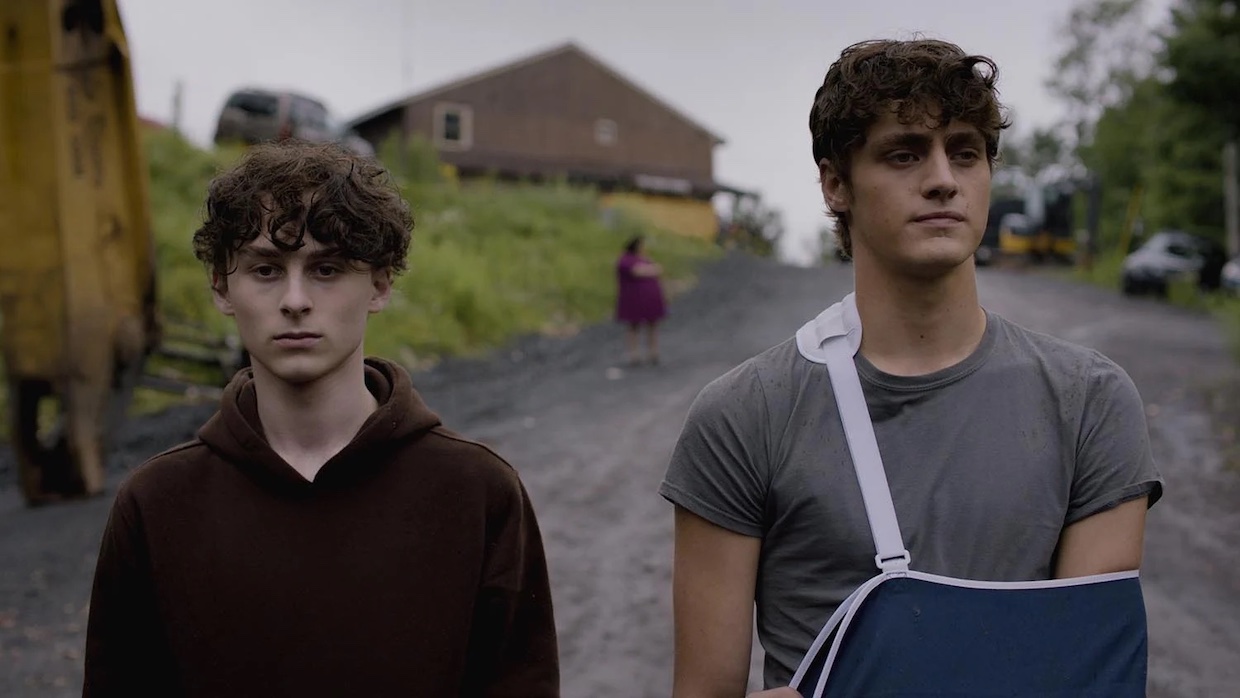 A boy with cropped curly hair and a brown hoodie stands next to his older brother, who's a bit taller and wears a gray t-shirt with his arm in a sling. In between them but behind in the distance is their mother, whose outline is blurred by her distance.