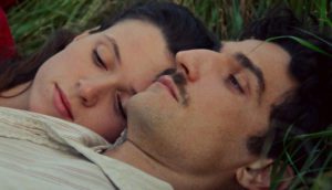 A young couple lie in the grass together. The woman rests her head on the man's shoulder.