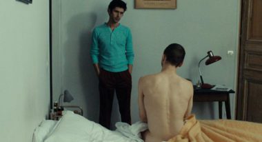 A man with a bright blue henley shirt and black pants stands in the corner of a bedroom and looks down at another man, who faces away from the camera and sits nude on the bed.