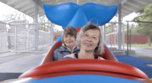 Two older women sit in an amusement park ride shaped like a whale, only the tail is visible behind them as they smile.
